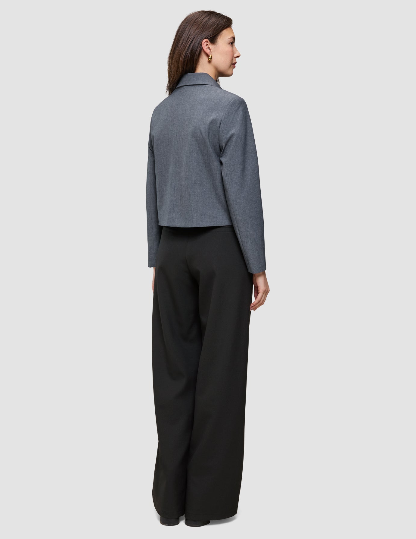 Kvinne sett bakfra, iført en Essential Short Zip Jacket i mørkegrå melange og svarte bukser med vide ben.